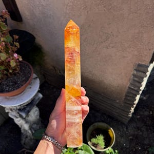 May include: A tall, rectangular citrine crystal point with a pointed top. The crystal has a gradient of colors, ranging from a light yellow to a deep orange. The crystal is held in a hand, with a bracelet visible on the wrist.