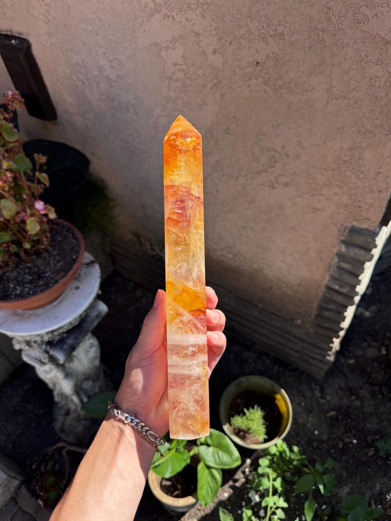 May include: A tall, rectangular citrine crystal point with a pointed top. The crystal has a gradient of colors, ranging from a light yellow to a deep orange. The crystal is held in a hand, with a bracelet visible on the wrist.