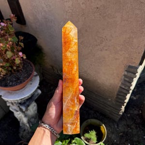 May include: A tall, rectangular, orange-yellow crystal obelisk. The crystal has a faceted top and a textured, translucent appearance. The obelisk is held in a hand, with a garden and building in the background. The crystal is approximately 8 inches tall.
