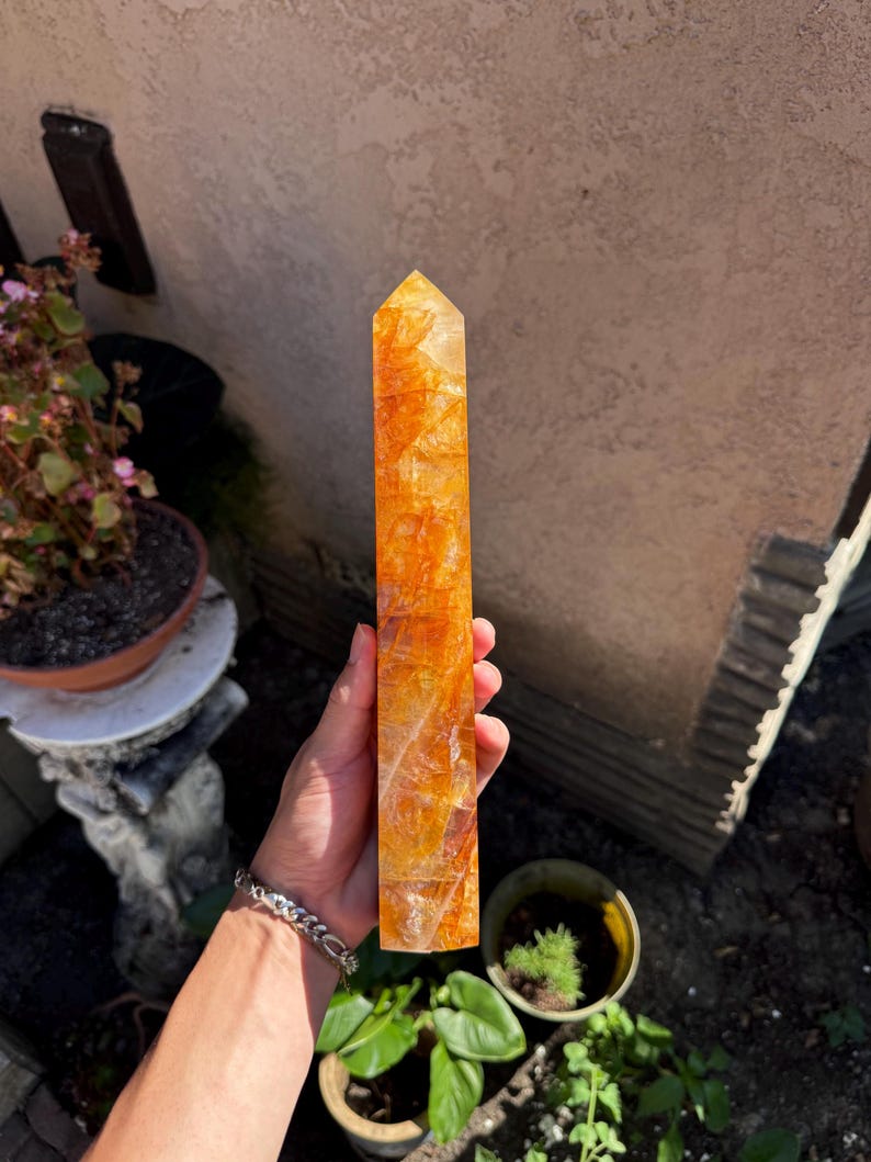 May include: A tall, rectangular, orange-yellow crystal obelisk. The crystal has a faceted top and a textured, translucent appearance. The obelisk is held in a hand, with a garden and building in the background. The crystal is approximately 8 inches tall.