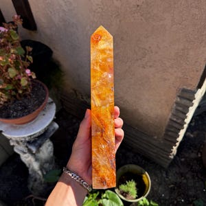May include: A tall, rectangular, orange-yellow crystal obelisk with a pointed top. The crystal has a translucent appearance with internal patterns. The obelisk is held in a hand, with a silver bracelet visible on the wrist. The background includes potted plants and a wall.