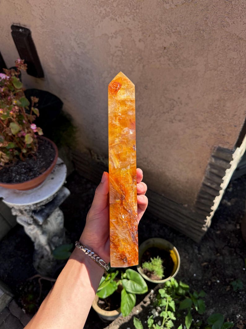 May include: A tall, rectangular, orange-yellow crystal obelisk with a pointed top. The crystal has a translucent appearance with internal patterns. The obelisk is held in a hand, with a silver bracelet visible on the wrist. The background includes potted plants and a wall.