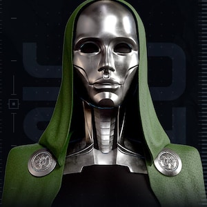 May include: A metallic silver mask with a smooth finish and a septum ring detail, worn under a textured green hood. The neck piece features segmented silver plating, and the hood is secured at the shoulders with two circular silver brooches.