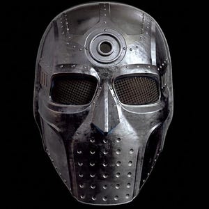 Puede incluir: Una máscara facial completa de metal plateado con construcción remachada, un emblema circular en la frente y cubiertas de malla oscura para los ojos. La sección inferior incluye un patrón perforado y la superficie tiene un acabado pulido.