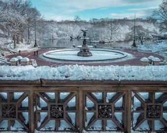Pode incluir: Cena de inverno de um parque coberto de neve com uma fonte no centro. A fonte tem uma estátua no topo e é rodeada por uma passarela circular. Árvores e edifícios ao fundo estão cobertos de neve.