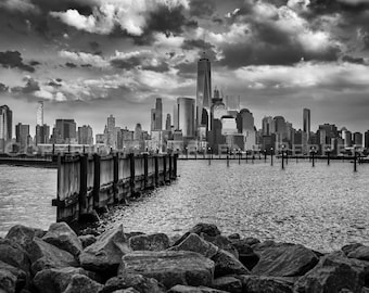 May include: Black and white photograph of the New York City skyline, featuring the One World Trade Center. The image includes a wooden pier, water, and a foreground of rocks under a cloudy sky. The overall tone is dramatic.