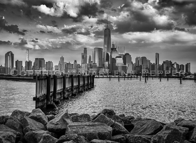 Pode incluir: Fotografia a preto e branco do horizonte da cidade de Nova Iorque, com o One World Trade Center. A imagem inclui um cais de madeira, &aacute;gua e rochas em primeiro plano, sob um c&eacute;u nublado e dram&aacute;tico.