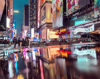 Pode incluir: Uma vista noturna da Times Square na cidade de Nova Iorque, com outdoors iluminados e reflexos em uma poça de água na rua.
