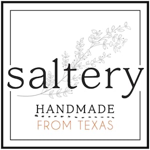 May include: A black and white logo with the word "saltery" in a stylized font. The logo is surrounded by a gray floral design. The text "HANDMADE FROM TEXAS" is below the logo.