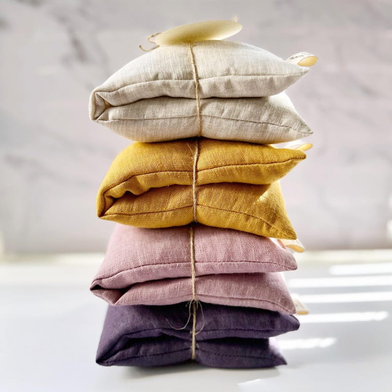 May include: A stack of four linen eye pillows in different colors: beige, yellow, pink, and purple. Each pillow is tied with twine.