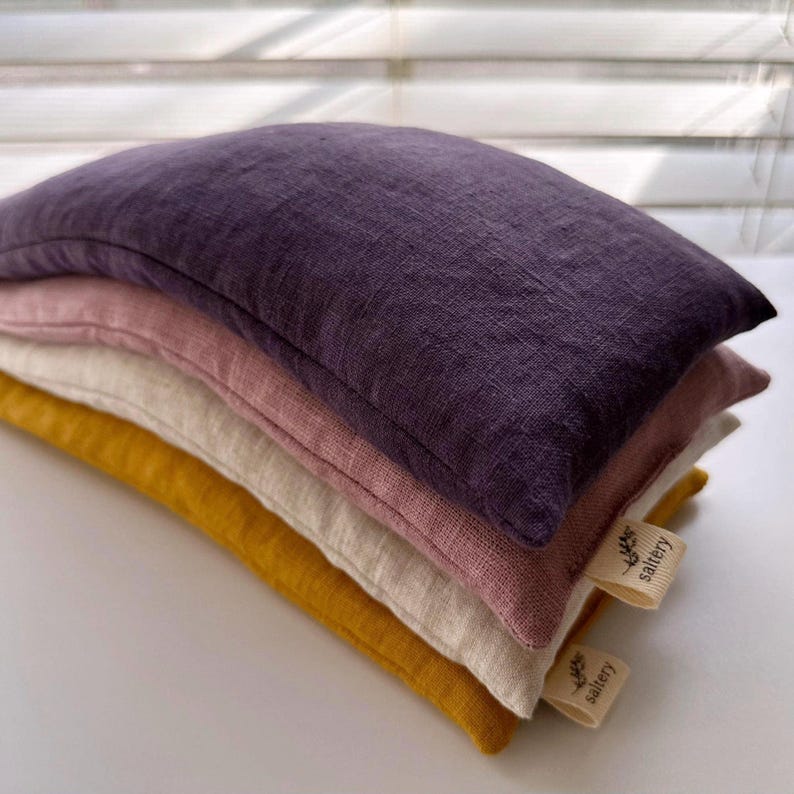 May include: Four linen eye pillows stacked on top of each other. The top pillow is purple, the next is pink, the next is beige, and the bottom pillow is yellow. Each pillow has a small label with the text "little saltery" on it.