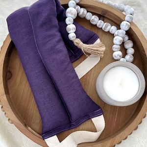 May include: A purple linen eye pillow with a white cotton strap, resting on a wooden tray with white beads and a white candle.