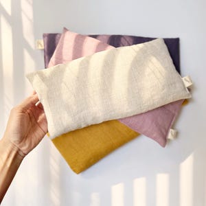 May include: Three linen eye pillows in different colors: beige, pink, and mustard yellow. The pillows are stacked on top of each other.