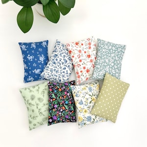 May include: A collection of small, rectangular pillows with various floral and patterned designs. The pillows feature a range of colors, including blue, red, green, and black, with white and yellow accents. The pillows are arranged on a white surface.