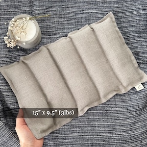 May include: A light gray linen weighted eye pillow measuring 15 inches by 9.5 inches and weighing 3 pounds. The pillow is made of four sections and has a label on the bottom right corner.