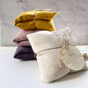 May include: Four heat therapy packs in different colors, including yellow, pink, purple, and beige. The beige pack is in the foreground and has a tag attached with instructions for using the pack. The tag reads "For Heat Therapy" and includes instructions for heating the pack in the microwave.