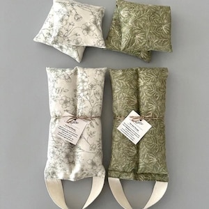 May include: Four floral-patterned heat packs in cream and green. Two square pillows and two long, rectangular packs with handles are visible. The packs are tied with twine and have tags. The floral patterns include delicate flower and leaf designs.