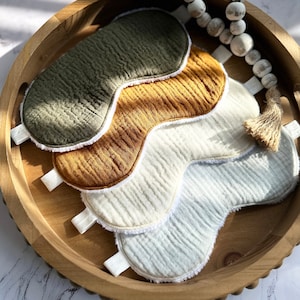 May include: Three eye masks made of soft, textured fabric. The masks are in shades of green, brown, and white. They are arranged in a stack on a wooden surface.