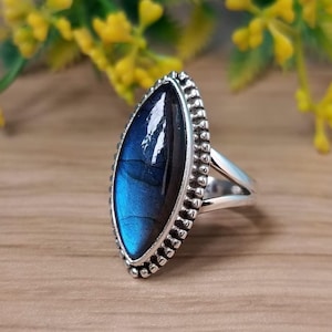 Puede incluir: Un anillo de plata con una piedra preciosa ovalada azul iridiscente. La piedra preciosa está engastada en un bisel con cuentas, y el anillo tiene un diseño de caña dividida. El anillo se muestra sobre una superficie de madera con flores amarillas en el fondo.
