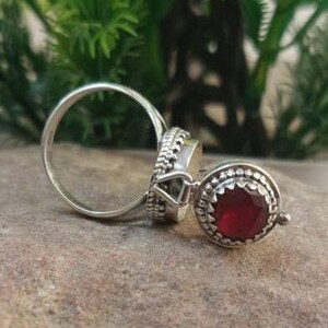 K&ouml;nnte beinhalten: Ein silberner Ring mit einem roten Edelstein, der in einer runden Fassung sitzt. Der Ring ist mit filigranen Details verziert und hat ein zartes Design.