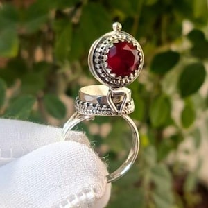 Könnte beinhalten: Ein silberner Ring mit einer kleinen, runden, klappbaren Box. Die Box ist geöffnet und zeigt einen roten Edelstein. Der Ring ist vor einem grünen, blättrigen Hintergrund platziert.