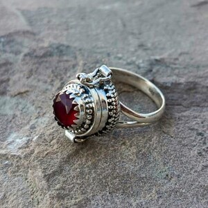 K&ouml;nnte beinhalten: Ein silberner Ring mit einem roten Edelstein und einem Scharnierfach. Der Ring liegt auf einer grauen, strukturierten Oberfl&auml;che.