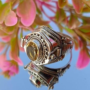 Pode incluir: Um anel de prata com um design detalhado e uma gema amarela. O anel é colocado sobre um fundo de flores cor-de-rosa.