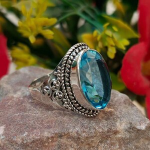 以下が含まれることがあります： 大きな楕円形の青い宝石が付いた銀の指輪。指輪は灰色の岩の上に置かれ、背景には赤い花が咲いています。