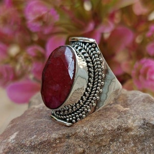 Peut inclure: Une bague en argent avec une grande pierre précieuse rouge de forme ovale sertie dans un décor en argent détaillé. La bague est posée sur une roche brune rugueuse.