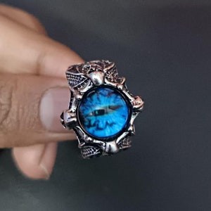 May include: A silver ring featuring a blue eye design. The ring has a detailed, intricate design surrounding the eye.