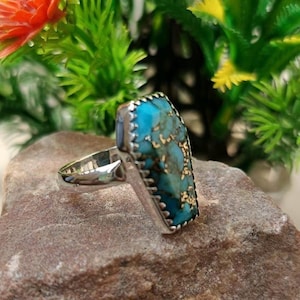 May include: A silver ring with a turquoise stone set in a geometric shape. The stone has a blue and green color with gold flecks.