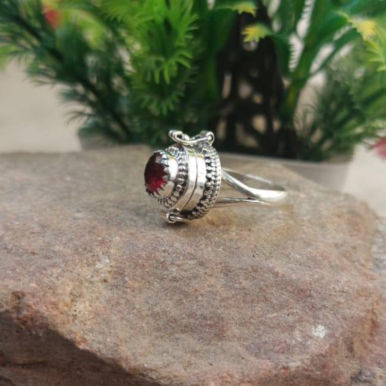 K&ouml;nnte beinhalten: Ein silberner Ring mit einem roten Edelstein und einem Scharnierfach. Der Ring liegt auf einem braunen Stein.