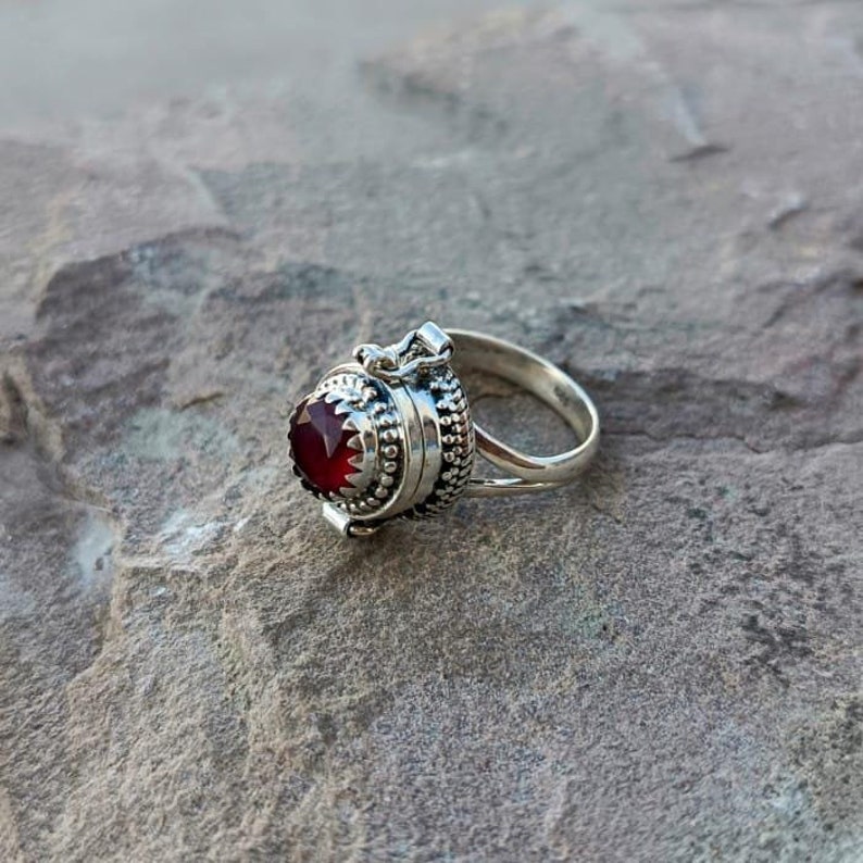 K&ouml;nnte beinhalten: Ein silberner Ring mit einem roten Edelstein und einem Scharnierfach. Der Ring liegt auf einer grauen, strukturierten Oberfl&auml;che.