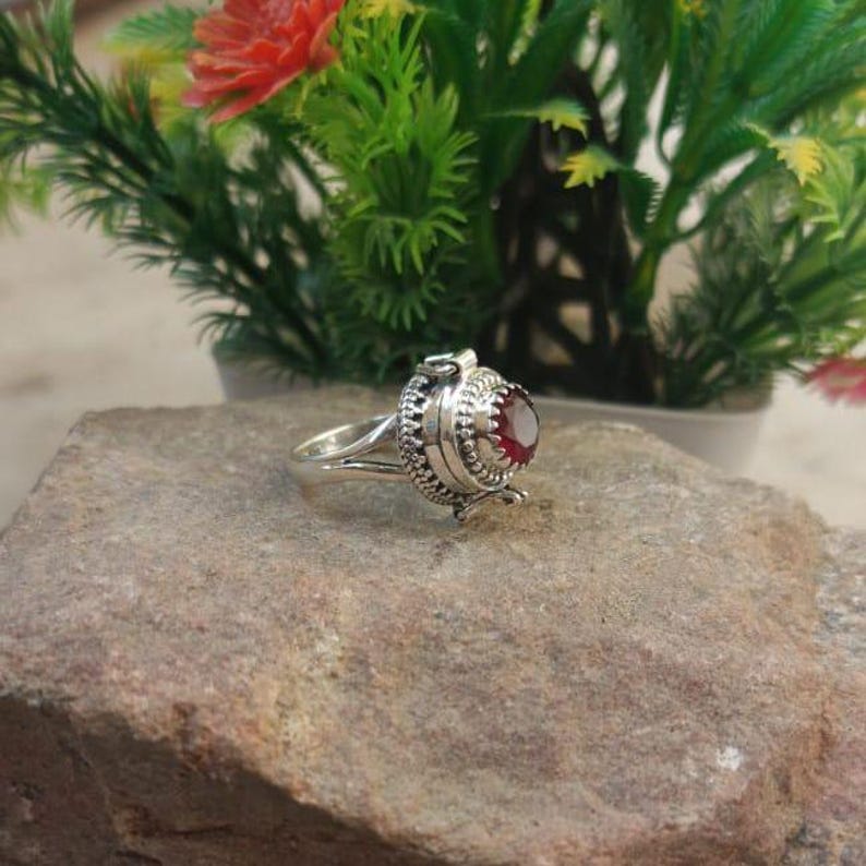 K&ouml;nnte beinhalten: Ein silberner Ring mit einem roten Edelstein und einem Scharnierfach. Der Ring liegt auf einem grauen Stein.