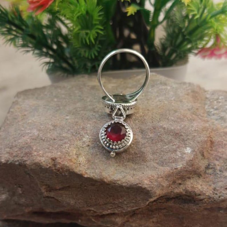 K&ouml;nnte beinhalten: Ein silberner Ring mit einem roten Edelstein, der herunterh&auml;ngt. Der Ring hat ein dekoratives Design und der Edelstein ist in einer silbernen Fassung gefasst.