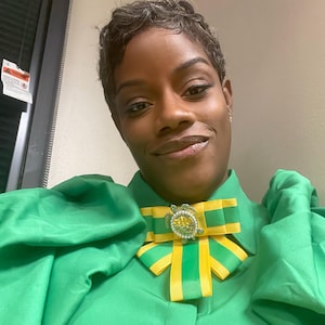 May include: A woman wearing a green blouse with a yellow and green bow tie featuring a jeweled turtle brooch.