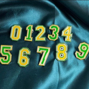 May include: Ten enamel pins featuring the numbers 0 through 9. The numbers are outlined in white and filled with yellow and green.
