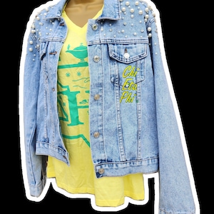 Peut inclure: Veste en jean bleu clair ornée de fausses perles, portée sur un t-shirt jaune à col en V. La veste présente des boutons argentés et le texte brodé "Chi Eta Phi" en jaune. Le t-shirt affiche un motif graphique vert.