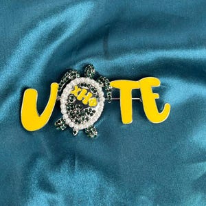 Può includere: Spilla in smalto giallo e bianco con la scritta "VOTE". La "O" è sostituita da un design a forma di tartaruga, impreziosito da strass verdi e perle bianche. La spilla è posta su uno sfondo di tessuto verde acqua. Accessorio decorativo.