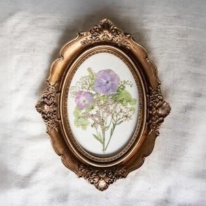 May include: An oval, antique gold-colored frame with ornate detailing surrounds a pressed flower arrangement. The arrangement features purple and pink flowers, green leaves, and white filler flowers, all set against a white background.