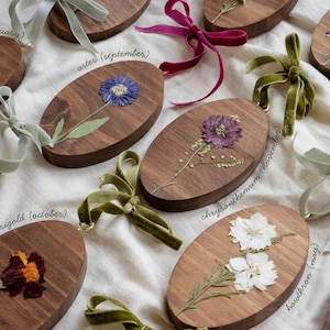May include: Oval wooden plaques with pressed flowers and velvet ribbons. Each plaque features a different flower, including violet, aster, marigold, chrysanthemum, and hawthorn. The names of the flowers and months are written on the plaques.