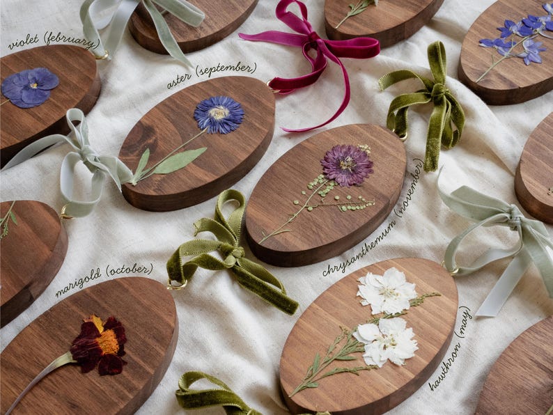 May include: Oval wooden plaques with pressed flowers and velvet ribbons. Each plaque features a different flower, including violet, aster, marigold, chrysanthemum, and hawthorn. The names of the flowers and months are written on the plaques.
