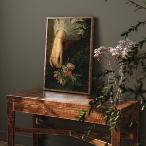 Pu&ograve; includere: Un dipinto incorniciato di una mano di donna che tiene un mazzo di fiori. I fiori sono un mix di rosso, rosa, bianco e giallo. La mano della donna indossa un polsino bianco. Lo sfondo &egrave; verde scuro e dorato.
