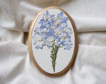 May include: Oval-shaped wall art featuring a bouquet of dried blue and white flowers. The flowers are mounted on a white canvas, framed by a gold-painted wooden border. The artwork is displayed against a white fabric backdrop.