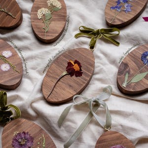May include: Wooden oval ornaments with pressed flowers and velvet ribbon hangers. Each ornament features a different flower, including cherry blossom, marigold, and aster, with the month of bloom labeled on the wood.