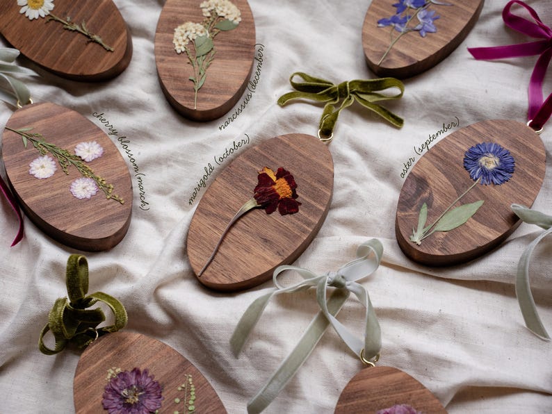 May include: Wooden oval ornaments with pressed flowers and velvet ribbon hangers. Each ornament features a different flower, including cherry blossom, marigold, and aster, with the month of bloom labeled on the wood.