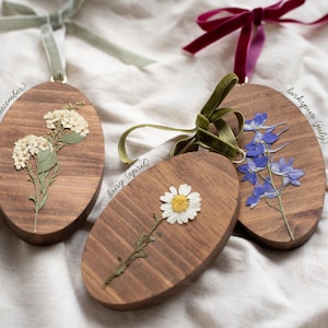 May include: Three wooden oval ornaments with dried flowers and attached ribbons. One features white flowers labeled "narcissus (december)", another a daisy labeled "daisy (april)", and the last blue flowers labeled "larkspur (july)".