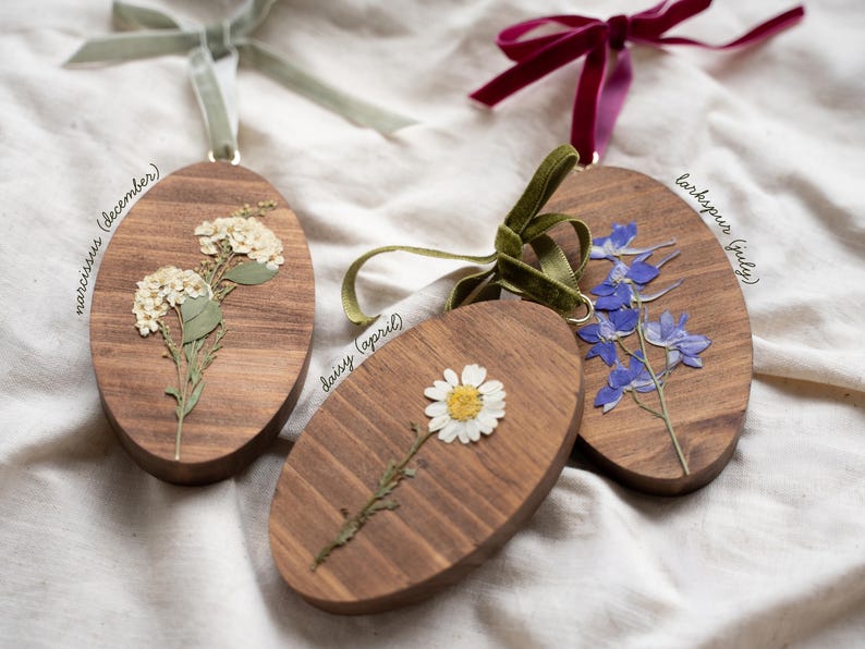 May include: Three wooden oval ornaments with dried flowers and attached ribbons. One features white flowers labeled "narcissus (december)", another a daisy labeled "daisy (april)", and the last blue flowers labeled "larkspur (july)".