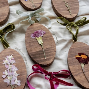 May include: Oval wooden ornaments with pressed flowers and velvet ribbon hangers. The ornaments feature various flowers, including carnations, gladiolus, and marigolds. Each ornament has a different colored ribbon.