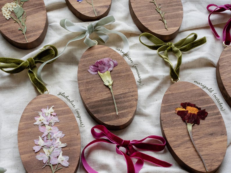 May include: Oval wooden ornaments with pressed flowers and velvet ribbon hangers. The ornaments feature various flowers, including carnations, gladiolus, and marigolds. Each ornament has a different colored ribbon.